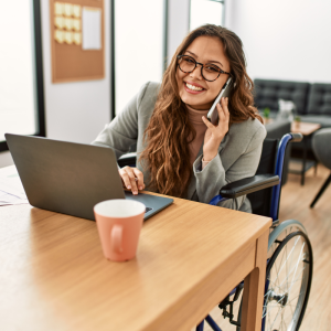 white woman in wheelchair talking on cell phone while working on a laptop at a table